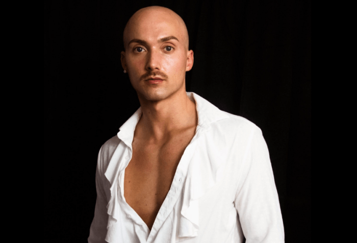 J. Logan Smilges, a white gender nonconforming trans person with a mustache, looks toward the camera without smiling. They are wearing a long-sleeve white shirt with ruffles down the middle and black pants with a checkered print. Photo credit: Darius Stokes
