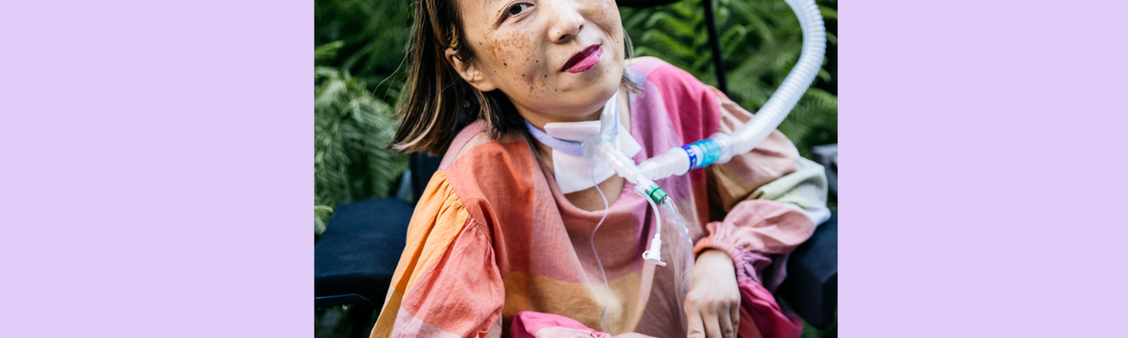 Graphic with a purple background. In the center is a photo of an Asian American woman in a wheelchair with a tracheostomy at her neck that has a tube connected to her ventilator. Her head is tilted to the left side, she is smiling, and she is wearing a magenta lip color. Her shirt is plaid with pale pink and other pastel colors and she is wearing pink pants. Behind her is a lot of greenery from bushes and trees at a park. Photo by @allisonbusch_photography