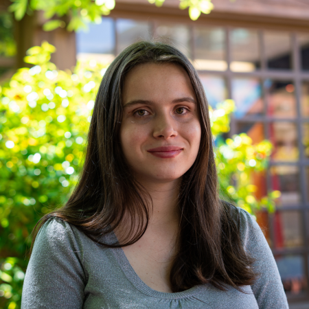 Julia Métraux, a white woman with dark brown hair smiling in a silver top. There are some bushes and windows behind her.