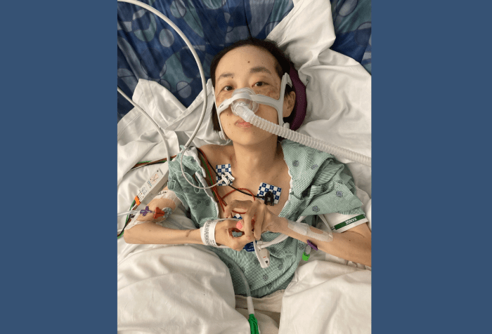 Graphic with a dark blue background. In the center is a photo of me in a hospital gown with tubes attached to my arms and face. Multiple pillows are underneath my arms and head. My hands are clasped together in the middle of my body. I have a serious look on my face.