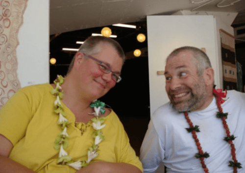 Petra and Neil are sitting side-by-side in their wheelchairs in or just outside a building in Honolulu. Petra, a smiling woman with short hair and glasses, is wearing a bright yellow top and wearing a lei as she gazes at Neil, to her left. Neil, a man with short hair and a beard, both dark with some gray mixed in, is wearing a white shirt and a lei, and gazing mischievously toward Petra. Courtesy of Petra Kuppers.