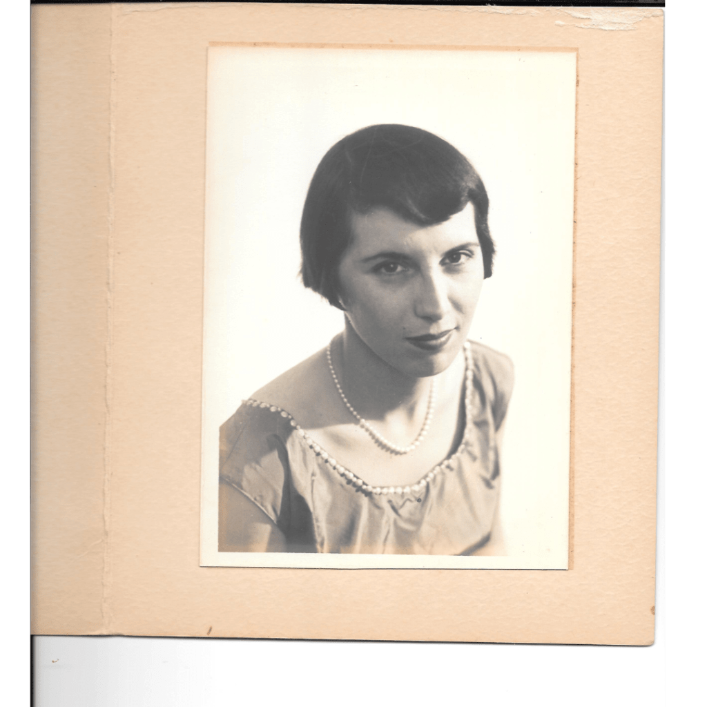 A formal portrait of my paternal grandmother. She wears a short sleeved dress with a square collar that is lined with beads, and she has a pearl necklace on. Her hair is in the beginnings of a short bob, as if she recently began growing it out. Her expression looks warm, but also formal. She has dark brown hair, dark brown eyes, and strong features, strong cheekbones.