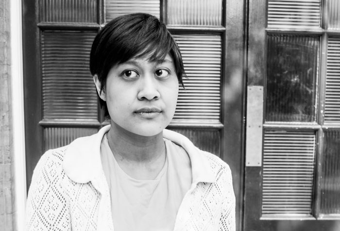 Black-and-white photo of an Indonesian woman in a cardigan and dress, with a door as backdrop. Picture credit: Wasi Daniju.
