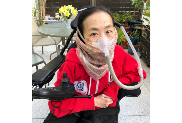Asian American woman in a power chair sitting at a patio with a red hoodie and a floral print handkerchief around her mouth. She’s wearing a mask over her nose attached to a gray tube.