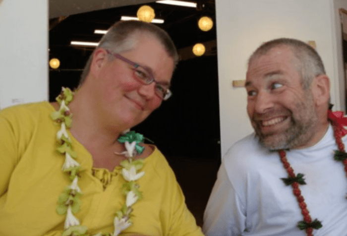 Petra and Neil are sitting side-by-side in their wheelchairs in or just outside a building in Honolulu. Petra, a smiling woman with short hair and glasses, is wearing a bright yellow top and wearing a lei as she gazes at Neil, to her left. Neil, a man with short hair and a beard, both dark with some gray mixed in, is wearing a white shirt and a lei, and gazing mischievously toward Petra. Courtesy of Petra Kuppers.