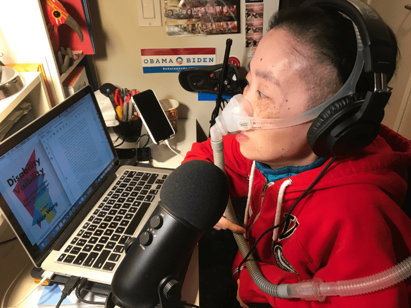 Photo of Alice Wong, an Asian American woman in a wheelchair at her desk with a laptop open. She is wearing a red hoodie and headphones over her ears. There is a mask over her nose attached to a ventilator. Next to the laptop is a microphone.