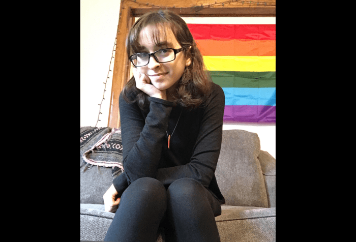 Graphic with a black background featuring a photo of Rachel, a white femme-presenting person sits in front of a pride flag. She has brown shoulder-length hair, and is wearing bronze earrings, black clothing, and black rimmed glasses. A little red piece of wood dangles from a thin silver necklace chain around her neck. She is sitting on a grey couch and rests her chin in one hand with one elbow on her knee. She grins slightly at the camera.