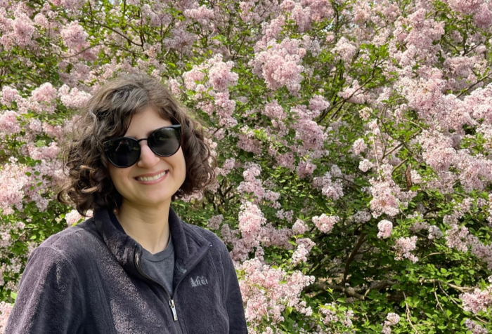 A photo of Raina, a smiling white woman with short curly brown hair. She is wearing sunglasses and a black jacket and is in front of a flowering lilac tree.