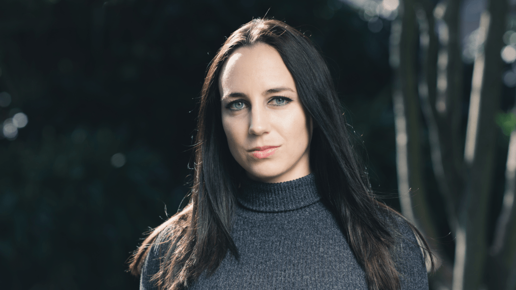 Heather Ringo, a white woman with long dark hair, wearing a gray turtleneck sweater, smiles wryly