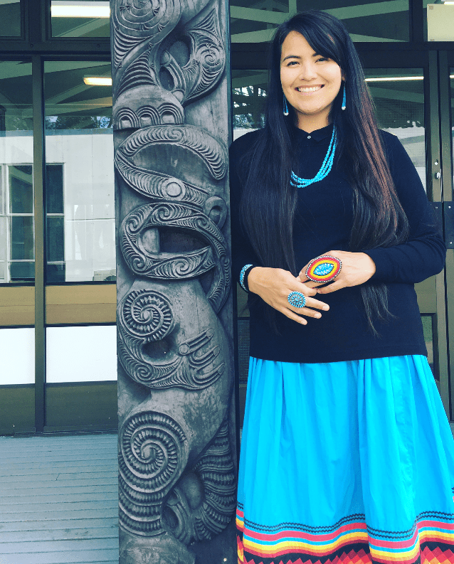 Photo of Sandra Yellowhorse. Sandra is smiling at the camera. She is outside and next to a Māori (Indigenous to New Zealand) timber carving of a taniwha (water spirit, powerful leader). She has long brown hair and is wearing a black sweater and turquoise skirt with black, red, orange and yellow woven bands. She is wearing turquoise earrings, a turquoise necklace and a large beaded ring that matches her skirt.