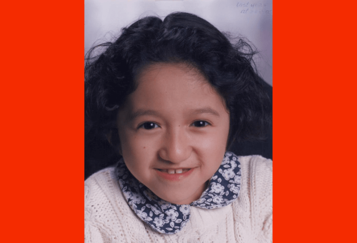 Graphic with a red background with a photo in the center of Sandy Ho in 5th or 6th grade. She has dark curly hair, is wearing a white sweater, and a blue and white flower collar. She is smiling at the camera.