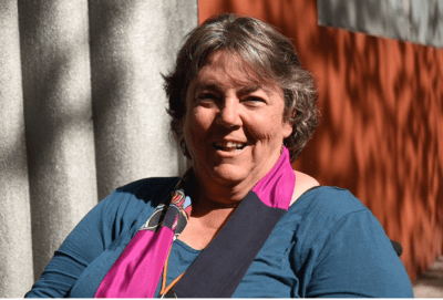 Photograph of Corbett, a large bodied white woman with greying hair. She wears a blue shirt and pink and blue scarf.