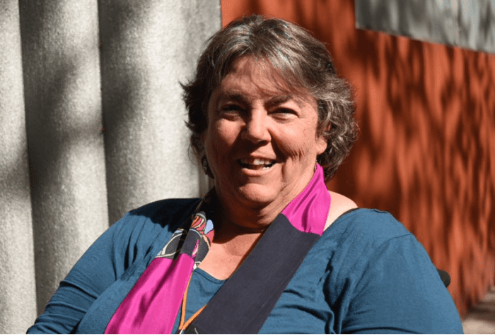 Photograph of Corbett, a large bodied white woman with greying hair. She wears a blue shirt and pink and blue scarf.