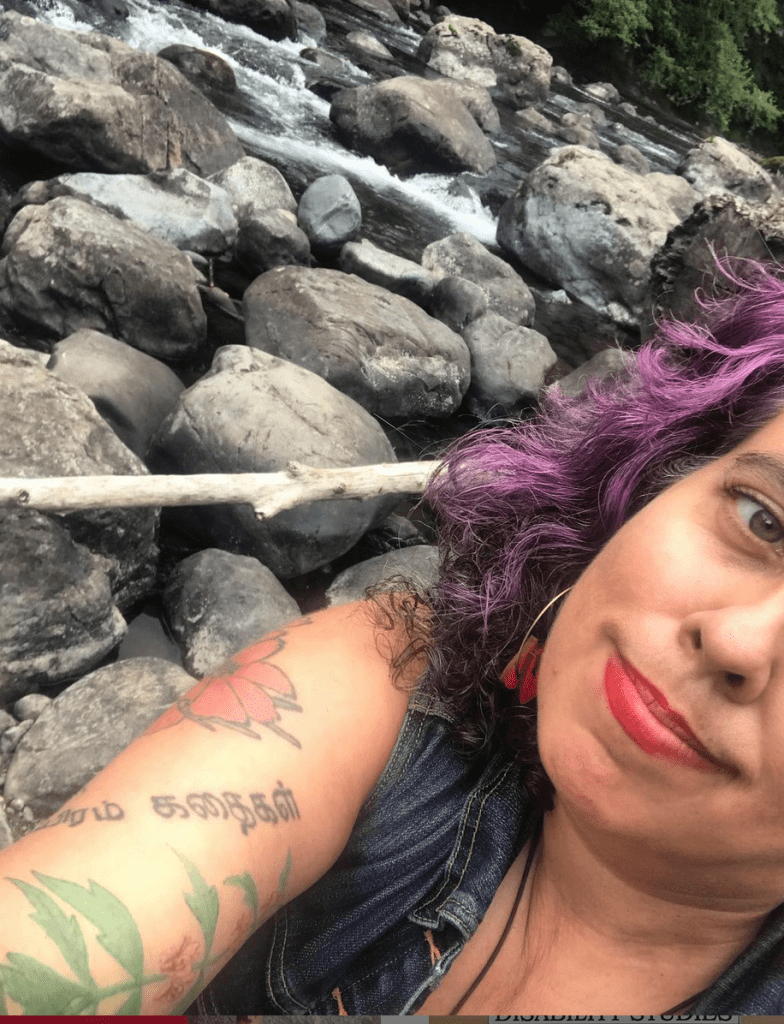 photo of a middle aged mixed race Sri Lankan, Irish and Romani femme, diagonially, in front of a wild river with lots of rocks. Leah has violet, brown and silver curly hair, peachy tan sand colored skin and red lipstick (Stila Stay All Day lipstick in Beso, aka "the lipstick AOC wears.") Their clothes are mostly not visible except for a blue denim vest. You can see their sleeve of tattoos, including cosmos flowers, a motherwort plant and letters in Tamil. They are smiling with bashful pride and satisfaction."