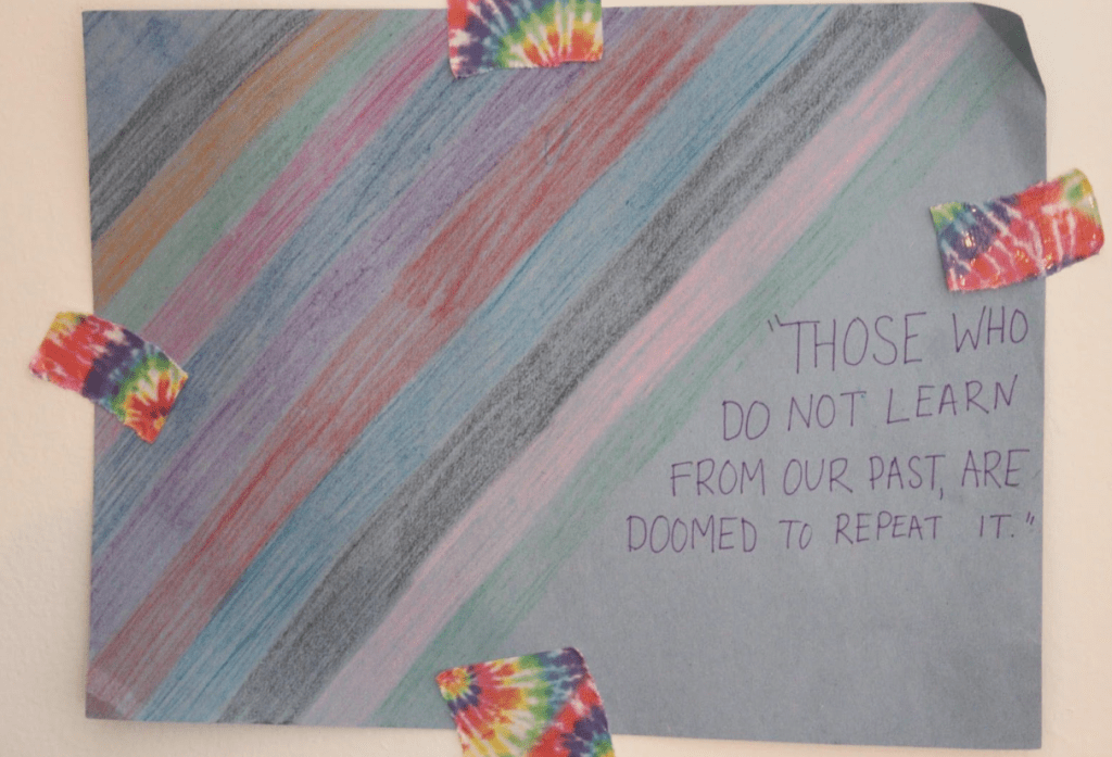 [THOSE WHO DO NOT LEARN FROM OUR PAST ARE DOOMED TO REPEAT IT (Purple font on blue paper) Rainbow stripes diagonal across the paper. It is attached to the wall with rainbow tie dye tape.]