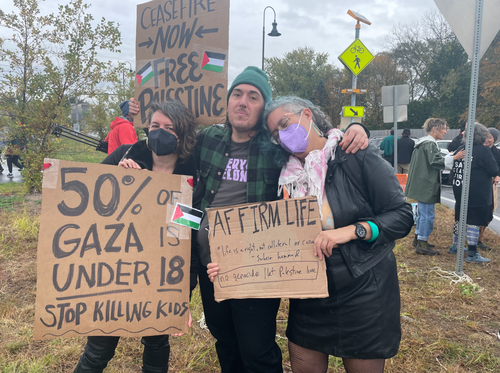 Image of Leah and two friends at a protest. One friend is a white, Greek/Irish femme person in her 40s with greying undercut brown hair wearing a black KN95 mask, they are holding a sign that says 50% OF GAZA IS UNDER 18 STOP KILLING KIDS. The second person is a Syrian masculine person wearing a green hat, a green and black flannel over a black t shirt with lilac lettering, holding a sign that reads CEASEFIRE NOW: FREE PALESTINE with two red black green and white Palestinian flags. Leah stands to the right leaning on their friend with their head on their shoulder, wearing a black leather jacket, black leather skirt, pink and white keffiyeh and lilac KN95 mask, holding a sign that reads, "Affirm life." Protestors can be seen in the background at a rotary in mid fall by a tree