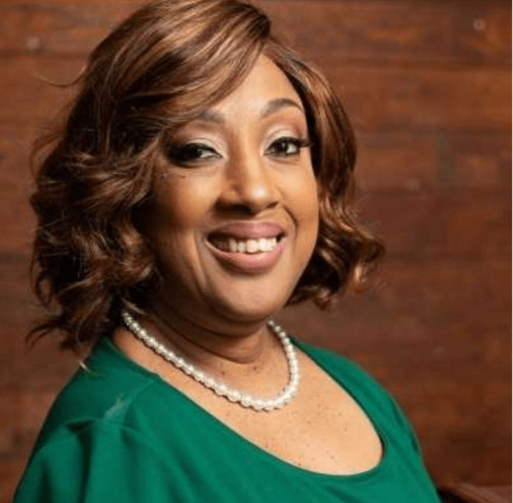 Foto de Rickii, mujer negra con pelo castaño medio rizado que sonríe a la cámara. Lleva collar de perlas y camisa verde.