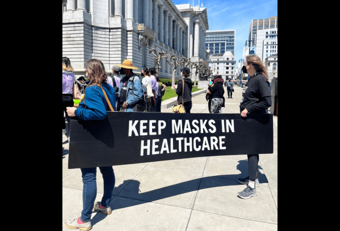 Graphic with a black background in the center is a photo of two people, facing to the left of the camera, are holding a black sign with white writing that says “Keep masks in healthcare”. In front of them is a crowd of people also faced to the left of the camera, at the corner of San Francisco's column enshrined City Hall.
