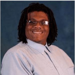 Smiling cis-male with brown complexion, glasses, dark brown eyes, and locs. He is wearing a light blue shirt