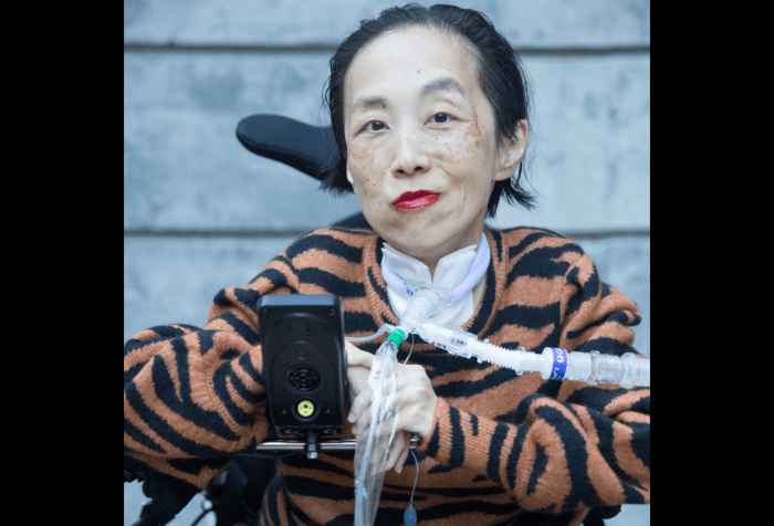 Graphic with a black background with a photo of Alice Wong, an Asian American disabled woman in a power chair. Behind her is a gray concrete wall. She is wearing a bold red lip color and an orange and black tiger striped sweater. A trach and ventilator tube is at her neck and shoulder. She is serving all the looks and giving a cheeky crooked smile. Photo credit: Eddie Hernandez Photography.