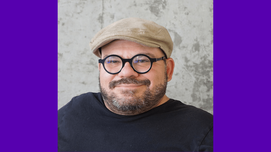 Graphic with a purple background, A photo of Joe’s head and shoulders on a gray background. He is a white man with a short cropped, dark beard and is wearing black rimmed round glasses, a black t-shirt, and tan colored flat cap.