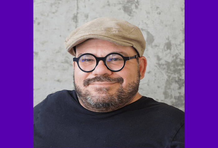 Graphic with a purple background, A photo of Joe’s head and shoulders on a gray background. He is a white man with a short cropped, dark beard and is wearing black rimmed round glasses, a black t-shirt, and tan colored flat cap.