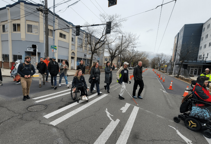 A group of a dozen adults, including some people using white canes, manual and power wheelchairs, cross a sidewalk in Seattle Washington. The sky is grey and everyone is bundled up in warm clothes. There are apartment buildings on either side of the street, and trees without leaves. 